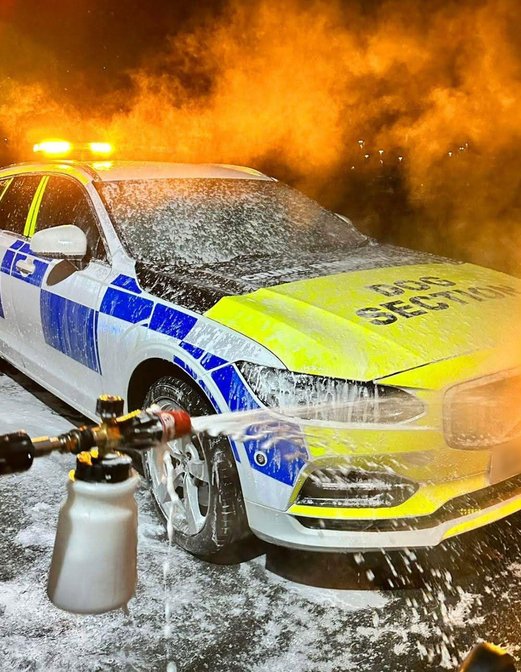 A police car with flashing lights is being washed with foam at night, with water spraying from a hose in the foreground.