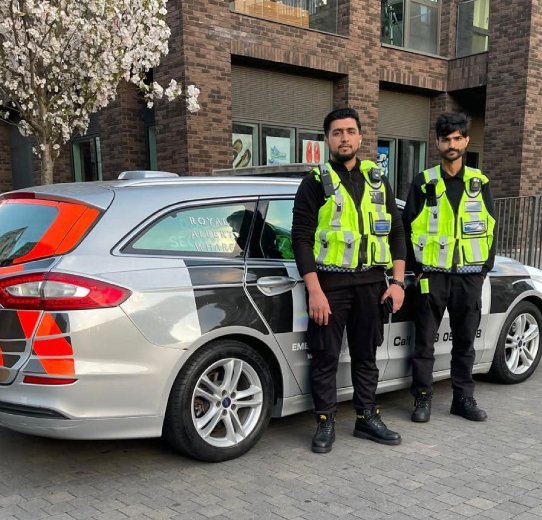 Two men in vests stand next to a police vehicle, likely involved in a professional or official interaction.